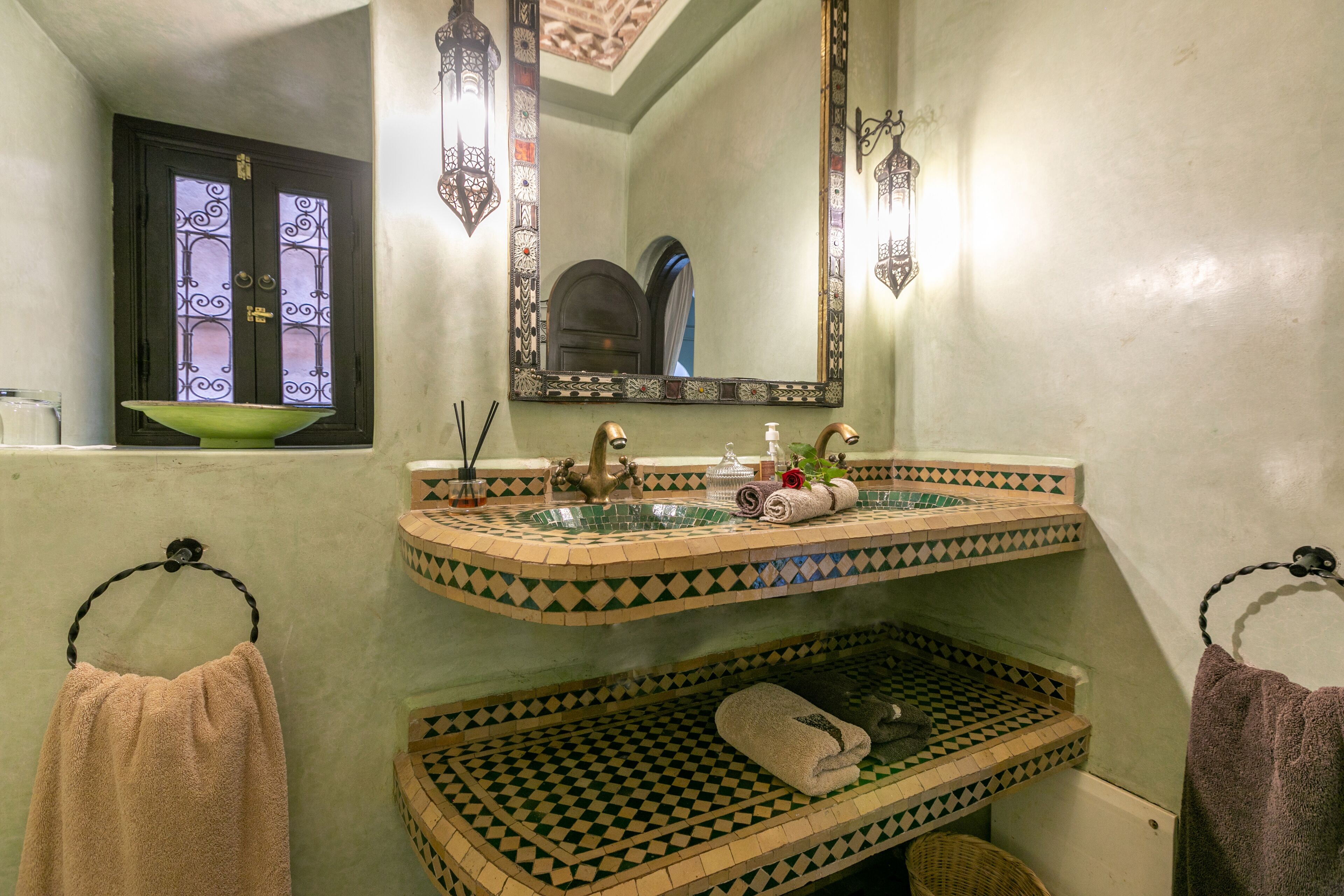 Une salle de bain avec un carrelage vert et blanc, un lavabo doté d’un receveur décoratif, un miroir encadré d’un cadre orné, et deux serviettes suspendues à des crochets.