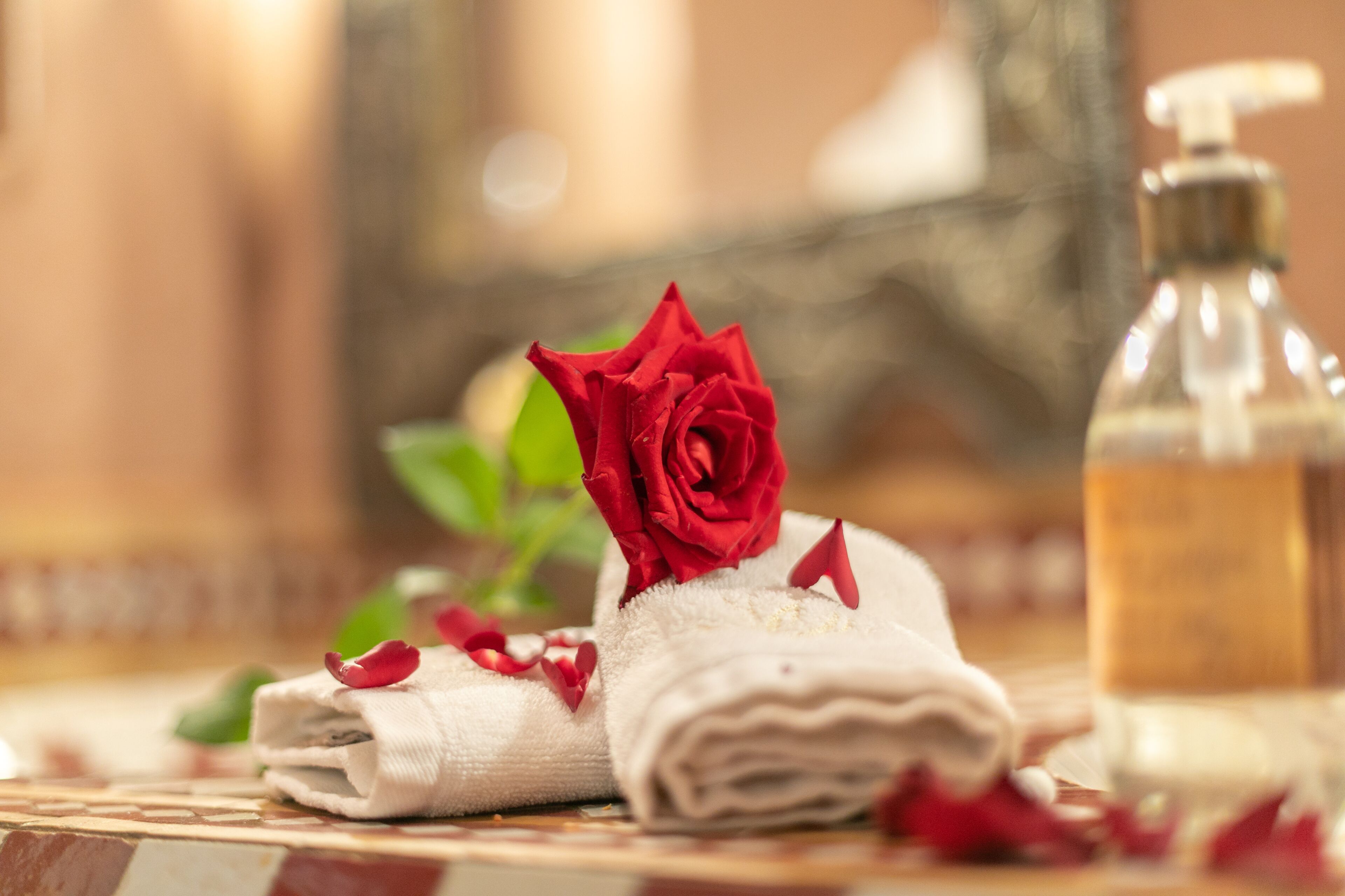 Et pænt foldet håndklæde med en rød rose og spredte rosenblade, en sæbe og et glas vand på en flisebelagt overflade.