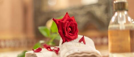 Et pænt foldet håndklæde med en rød rose og spredte rosenblade, en sæbe og et glas vand på en flisebelagt overflade.