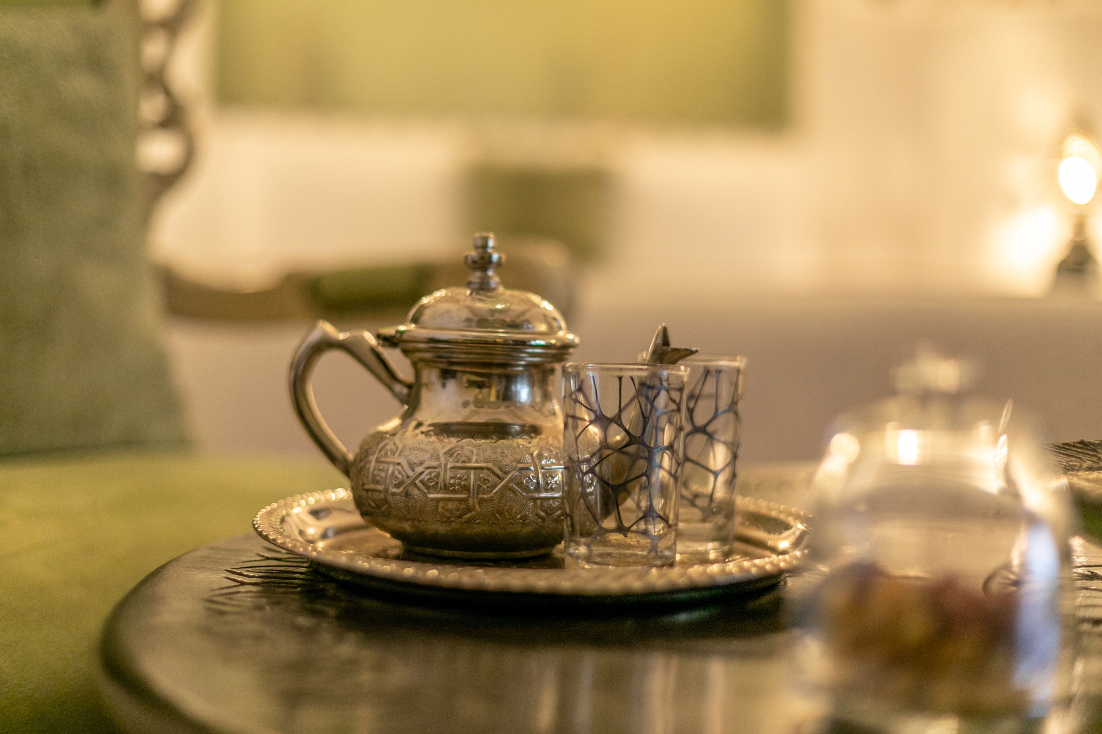 Une table avec une théière en argent, un verre orné d’un motif géométrique et un bocal en verre, le tout posé sur un plateau.