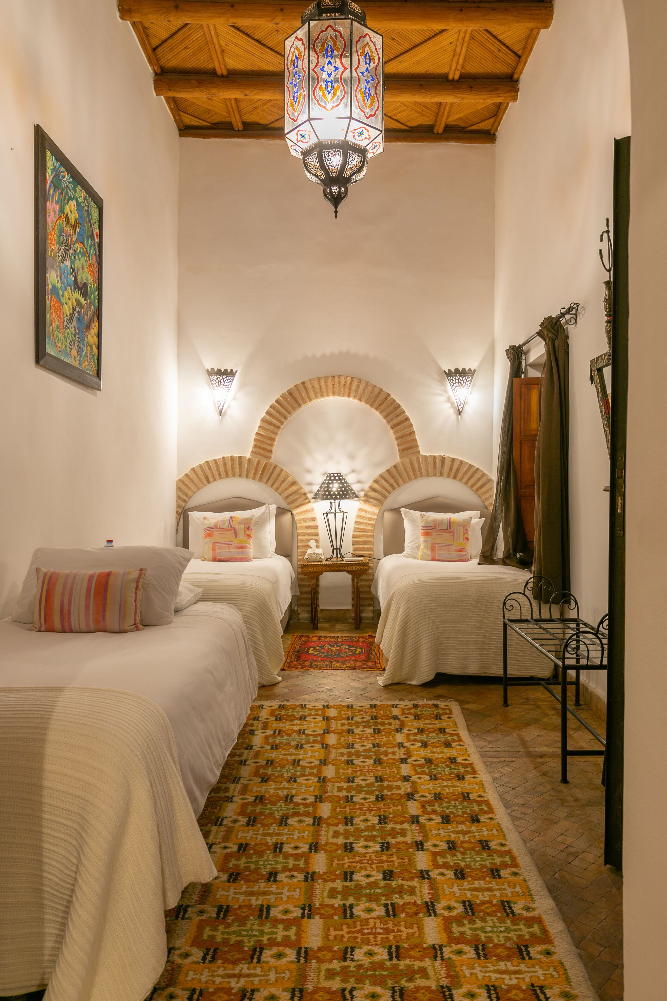 A room with two beds, a patterned rug, a chandelier, and a decorative wall arch.
