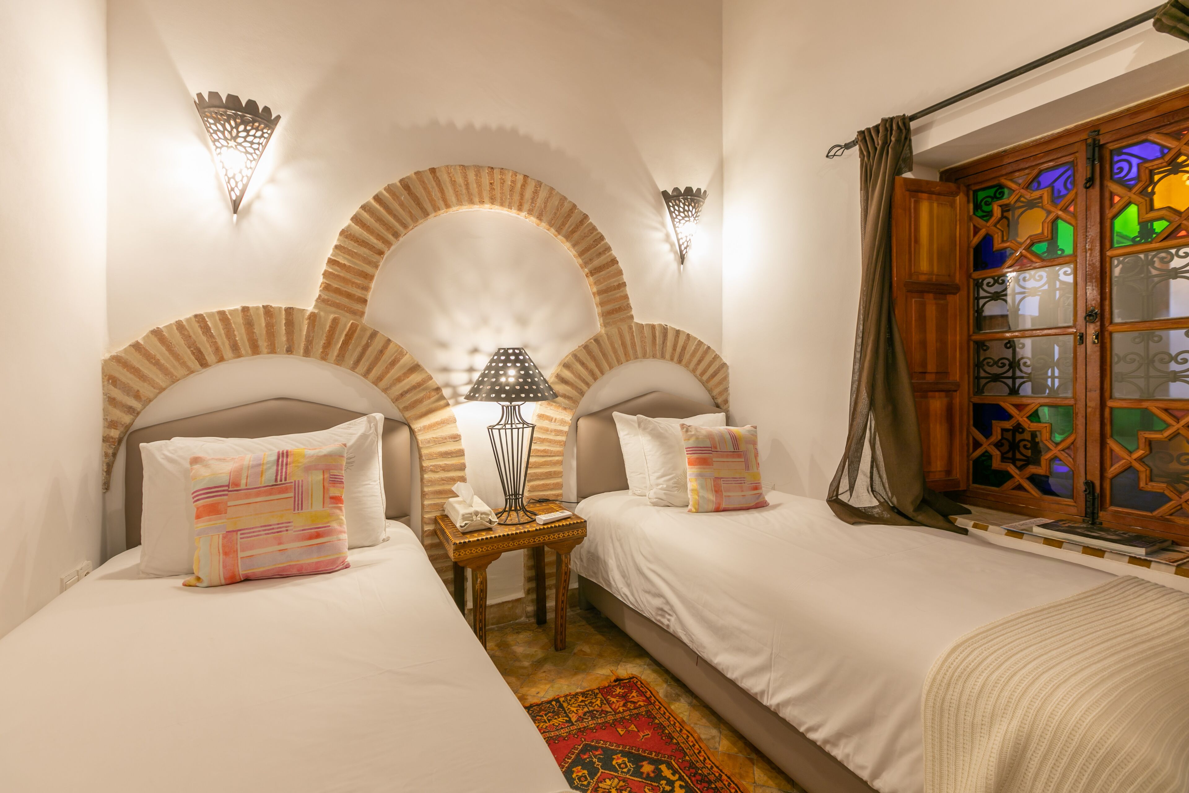 A room with two beds, a small table with a lamp, and a window with decorative glass.