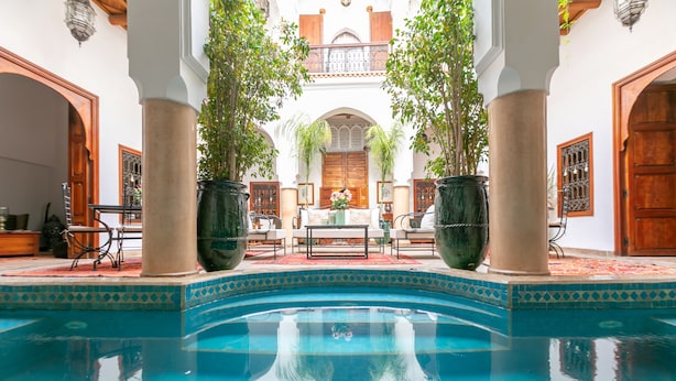 Zona de piscina con borde de azulejos, plantas en macetas y una mesa con sillas.