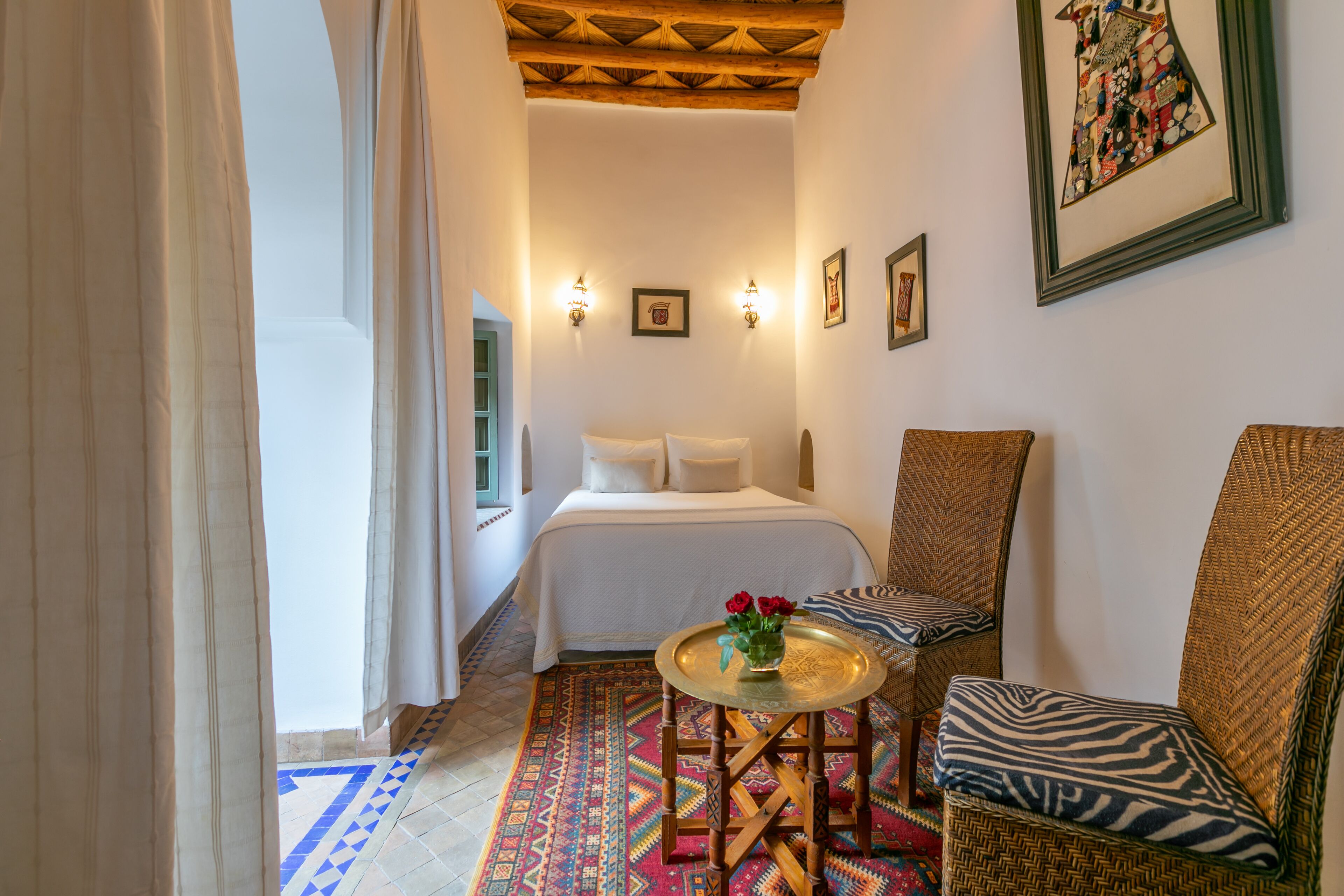 Une chambre à coucher avec un lit, deux lampes fixées au mur, une petite table ronde avec un vase et une chaise agrémentée d’un coussin à motifs.