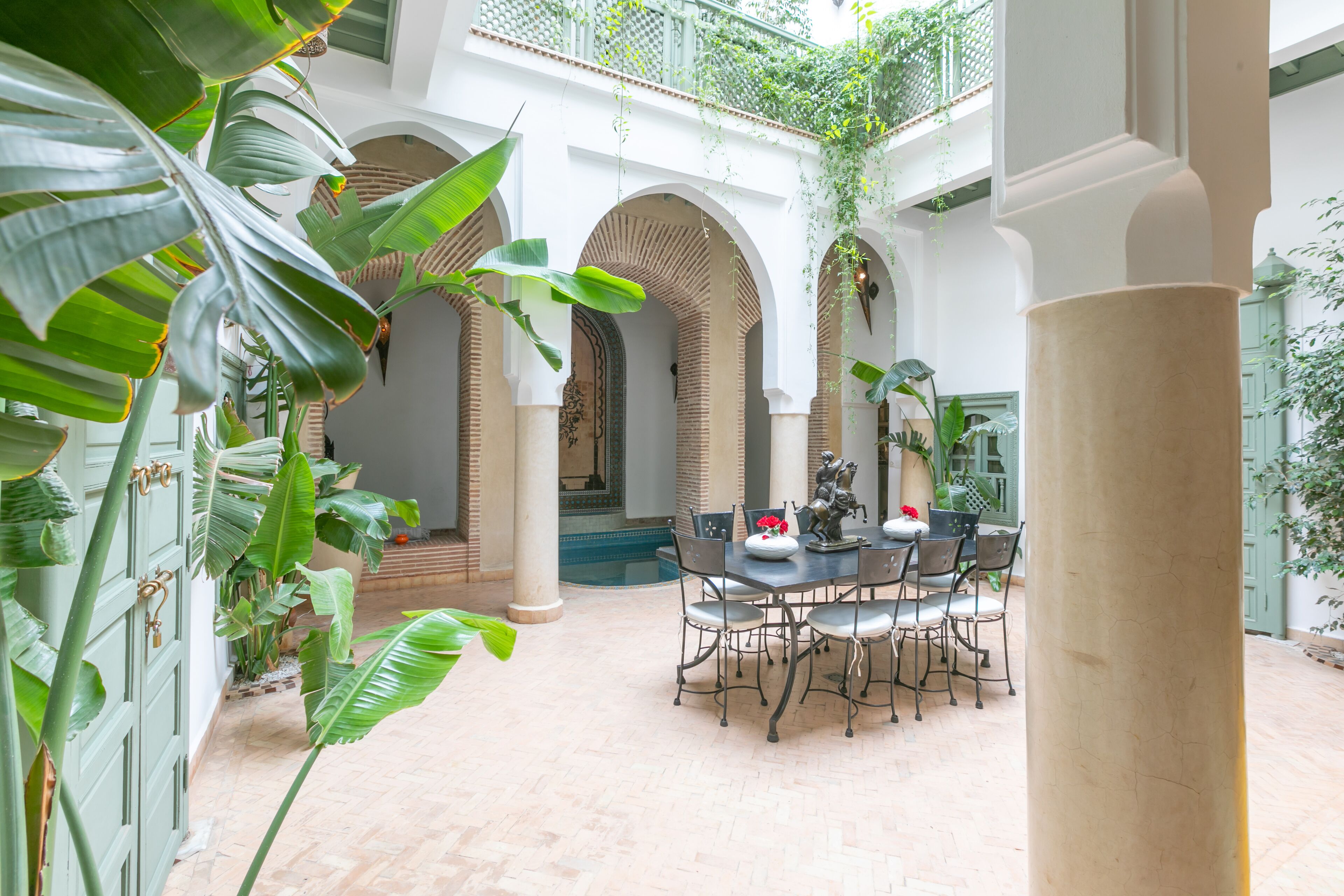 Une cour dotée d’une table ronde et de chaises, entourée d’arcades et de colonnes, avec une végétation luxuriante et une vue sur une piscine.