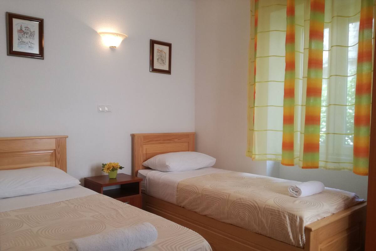 Une chambre d’hôtel avec deux lits, une table de chevet ornée d’un vase de fleurs et une fenêtre avec des rideaux colorés.