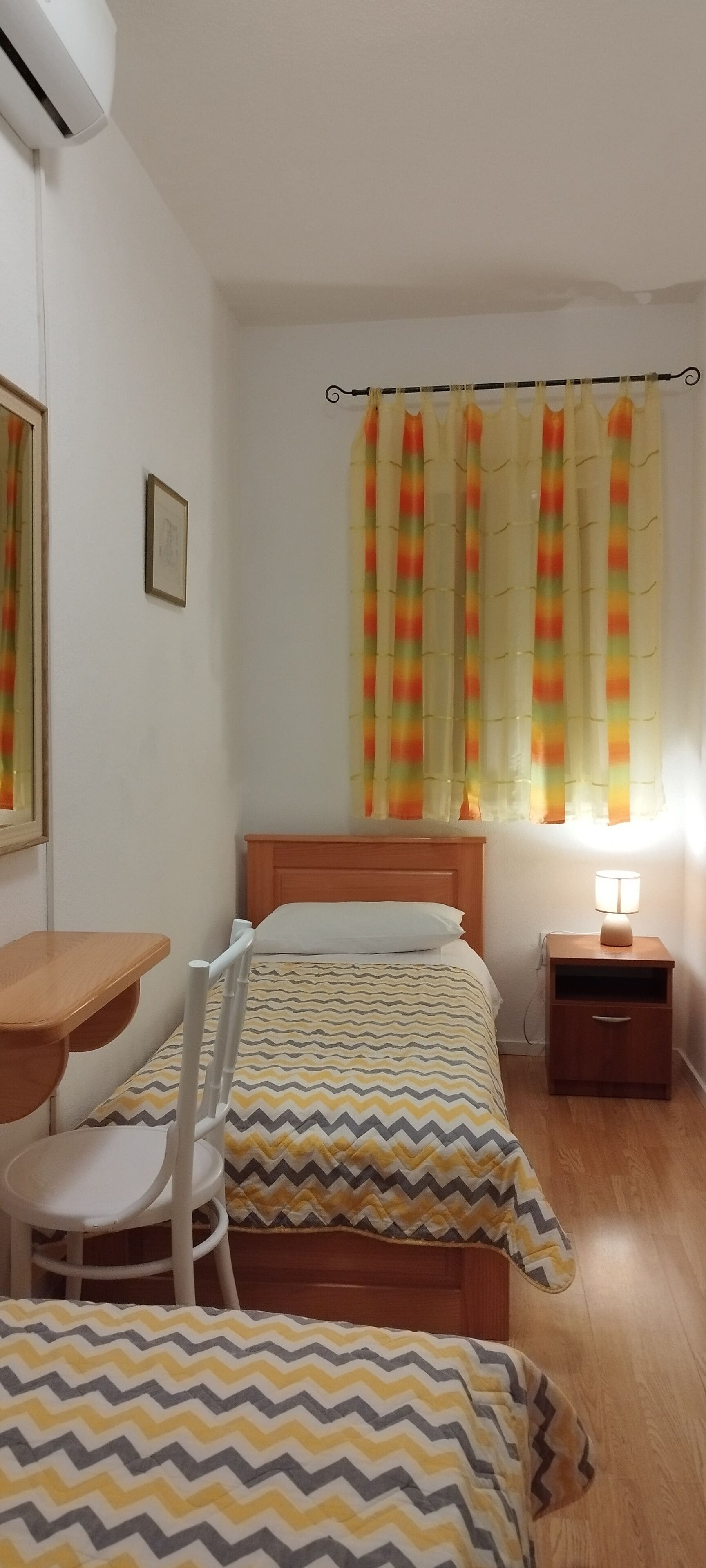 A bedroom with two beds, a desk, a chair, and a nightstand with a lamp.
