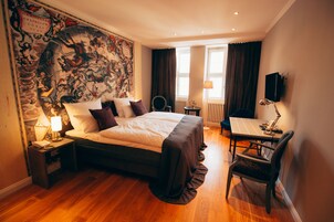 A hotel room with a large bed, a desk with a chair, a desk lamp, a nightstand with a lamp, and a large tapestry on the wall.