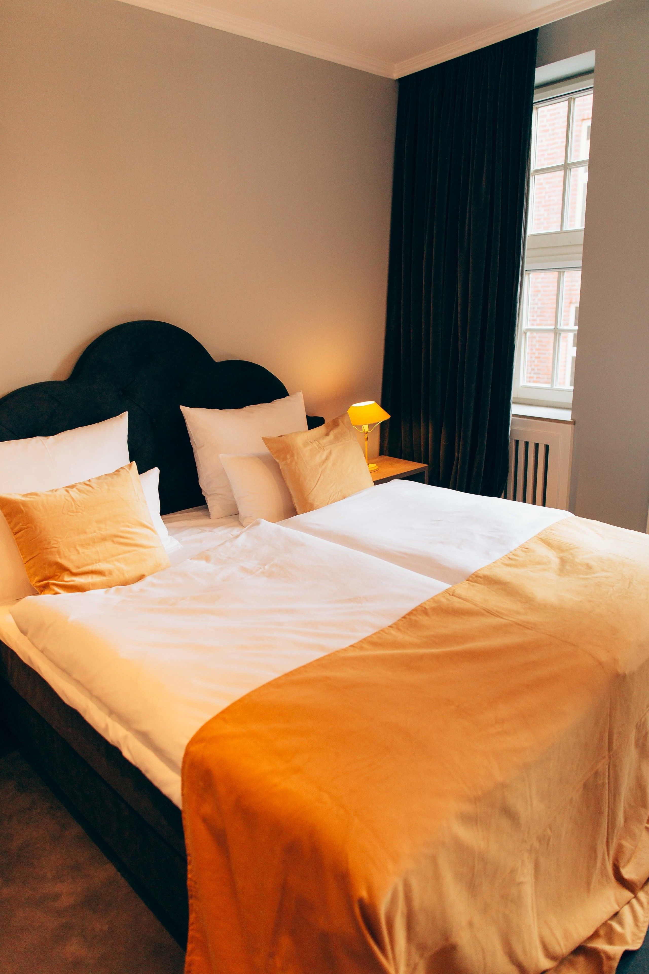 Una cama bien tendida con ropa de cama blanca y beige, un cabecero oscuro y una ventana con cortinas.