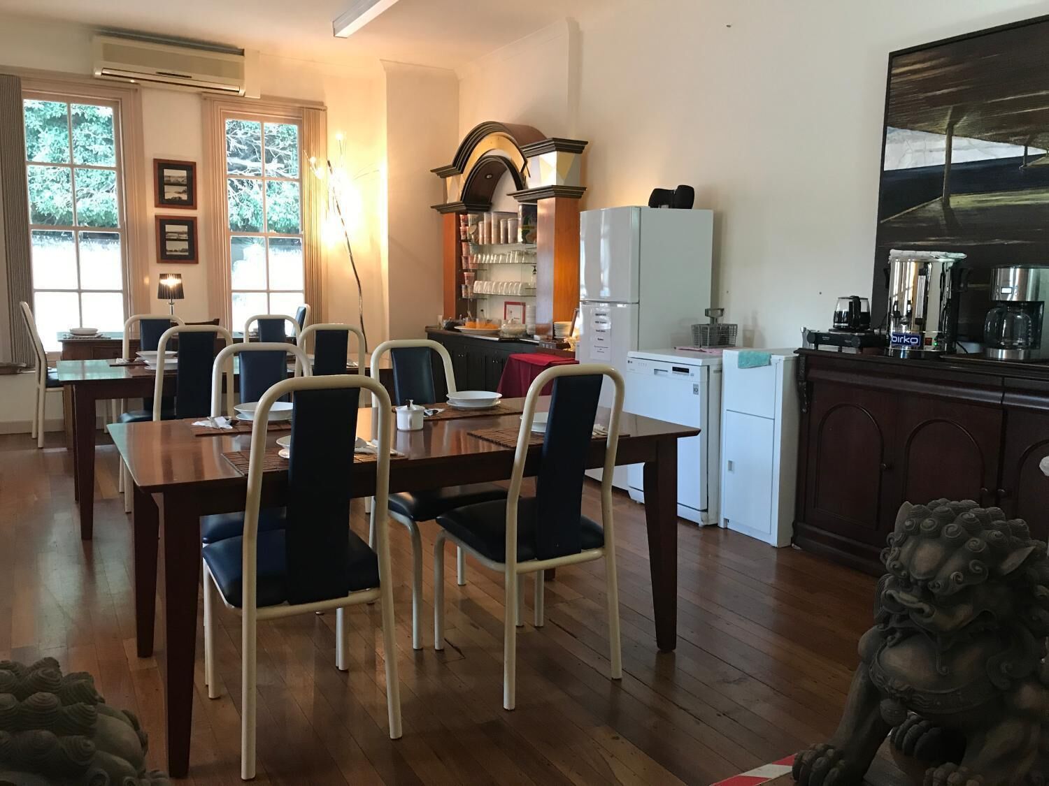 A dining area with a wooden table and chairs, a kitchen with a refrigerator and coffee machine, and a statue in the foreground.