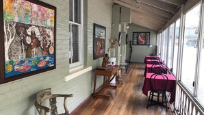 A covered outdoor seating area with wooden tables and chairs, artwork on the walls, and a view of the street.