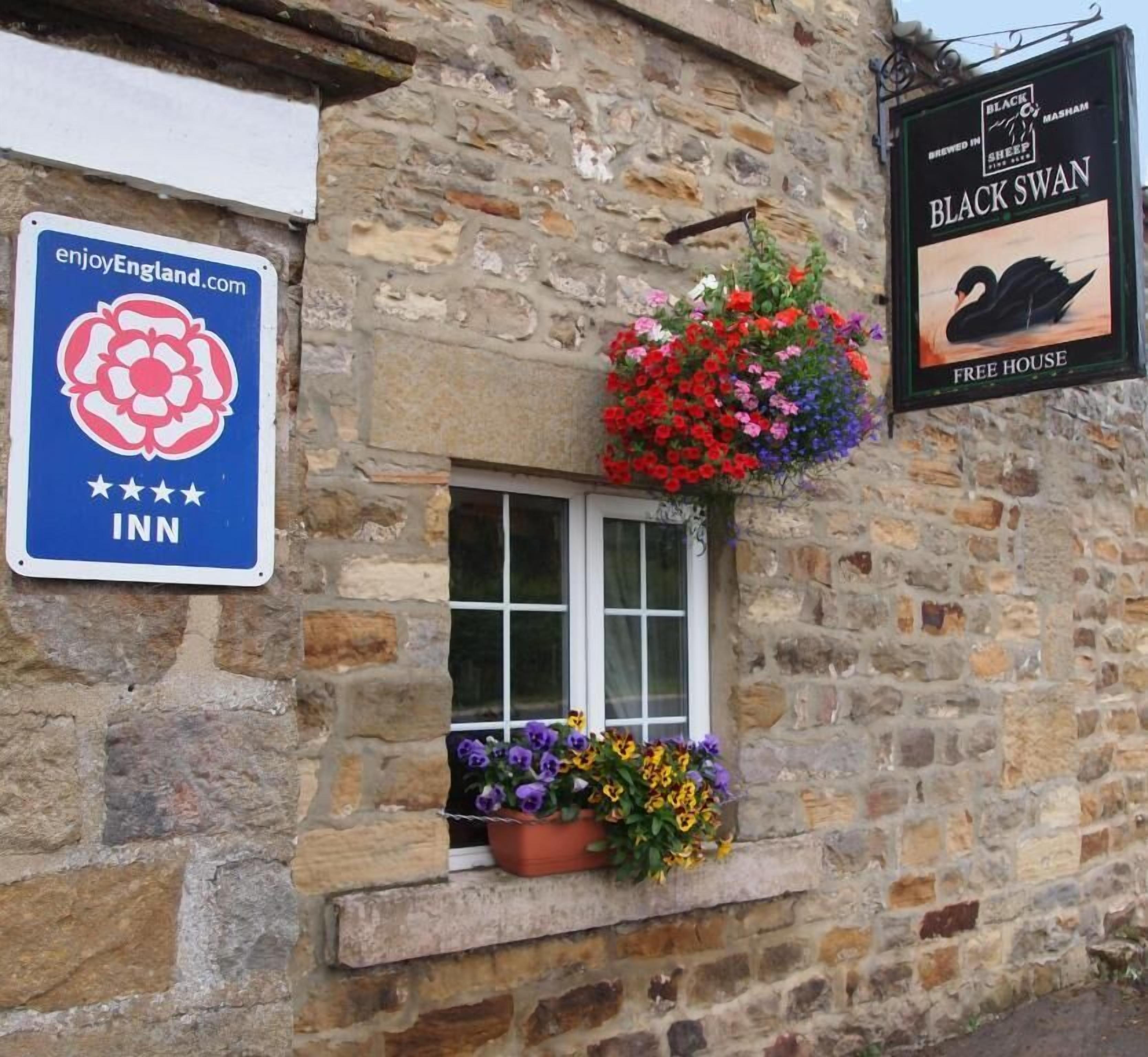 Un bâtiment en pierre avec une fenêtre ornée de fleurs colorées. On peut y voir des panneaux indiquant « Black Swan Free House » et « Enjoy England Inn ».