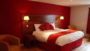 A bedroom with a wooden headboard, a large bed with red and white bedding, two bedside lamps, a nightstand, and a chair. - Black Swan Inn (Ripon)