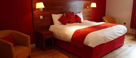 Quarto com cabeceira de madeira, uma cama grande com roupa de cama vermelha e branca, duas luminárias de cabeceira, uma mesa de cabeceira e uma cadeira.