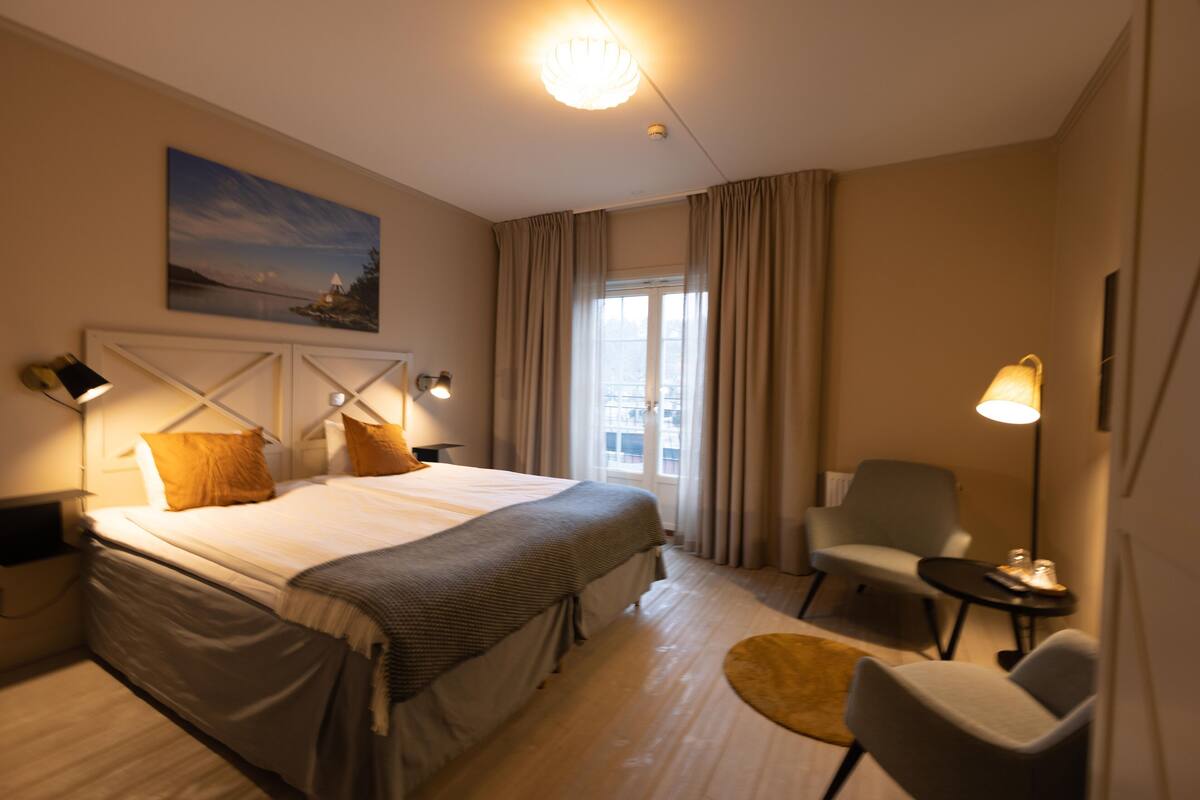 Una habitación de hotel con cama, una silla, una mesita, una lámpara, un cuadro en la pared y una ventana con cortinas.