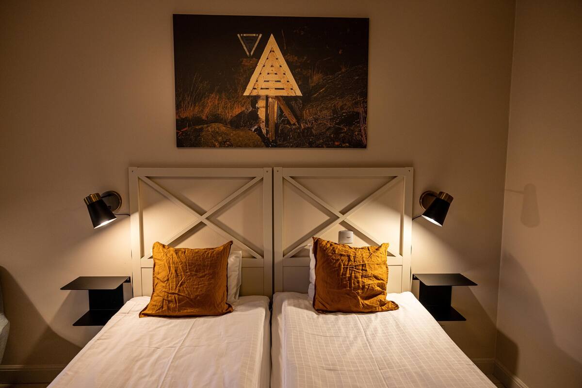 Un dormitorio con dos camas, cada una con una colcha blanca y una almohada marrón. Hay dos mesitas de noche con lámparas, y un cuadro de una estructura triangular cuelga sobre las camas.