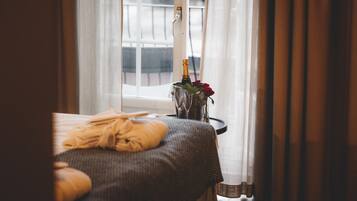 Un lit avec une couverture grise et une serviette jaune, un vase avec des fleurs et une bouteille sur une table de chevet, et une fenêtre avec des rideaux.
