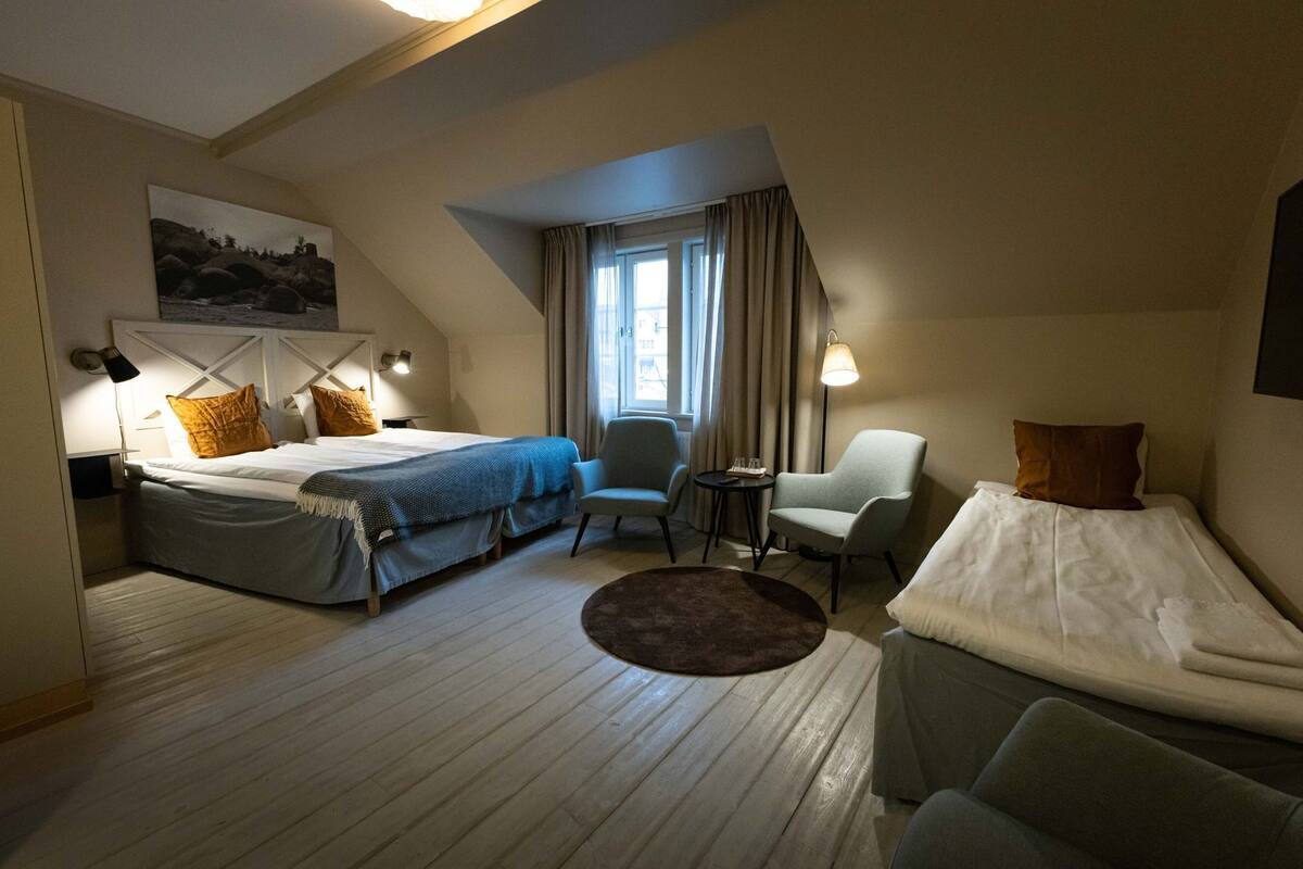 Habitación de hotel con dos camas, una mesita, dos sillas, una ventana con cortinas y un cuadro en la pared.