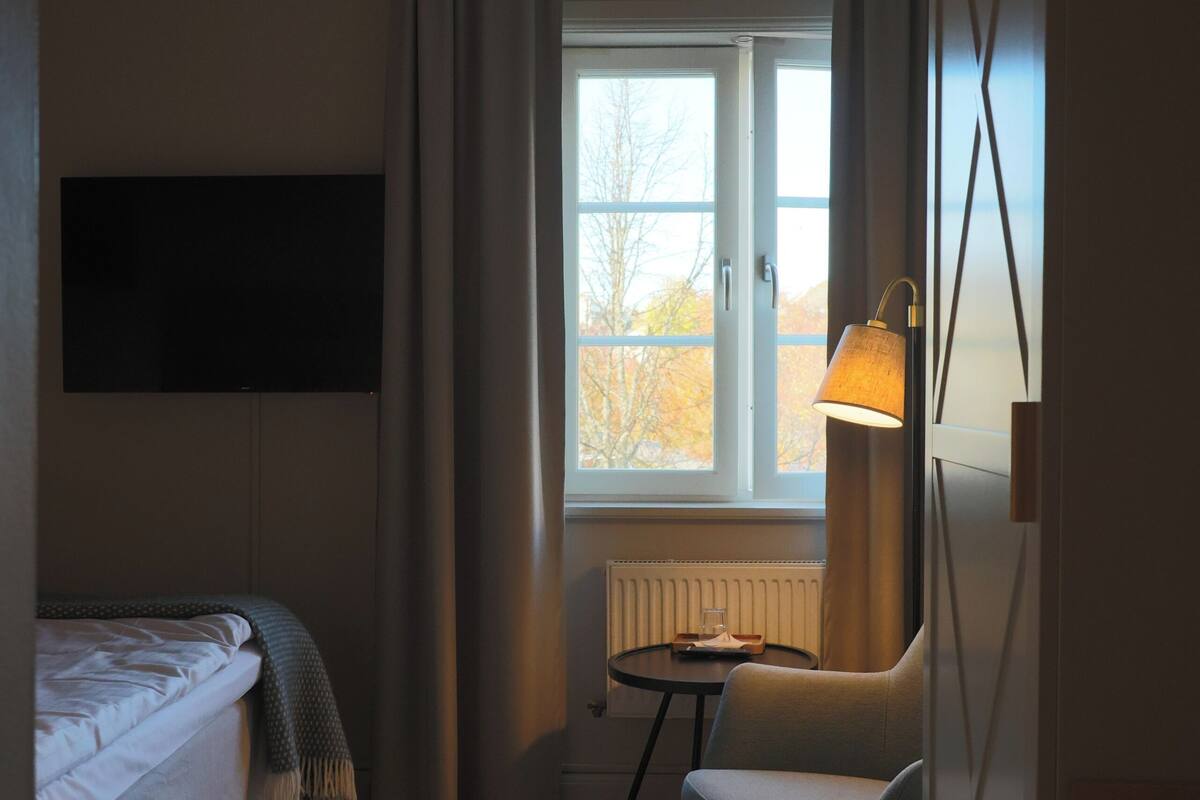 Habitación de hotel con cama, una silla, una mesita, lámpara, televisor montado en la pared y ventana con cortinas.