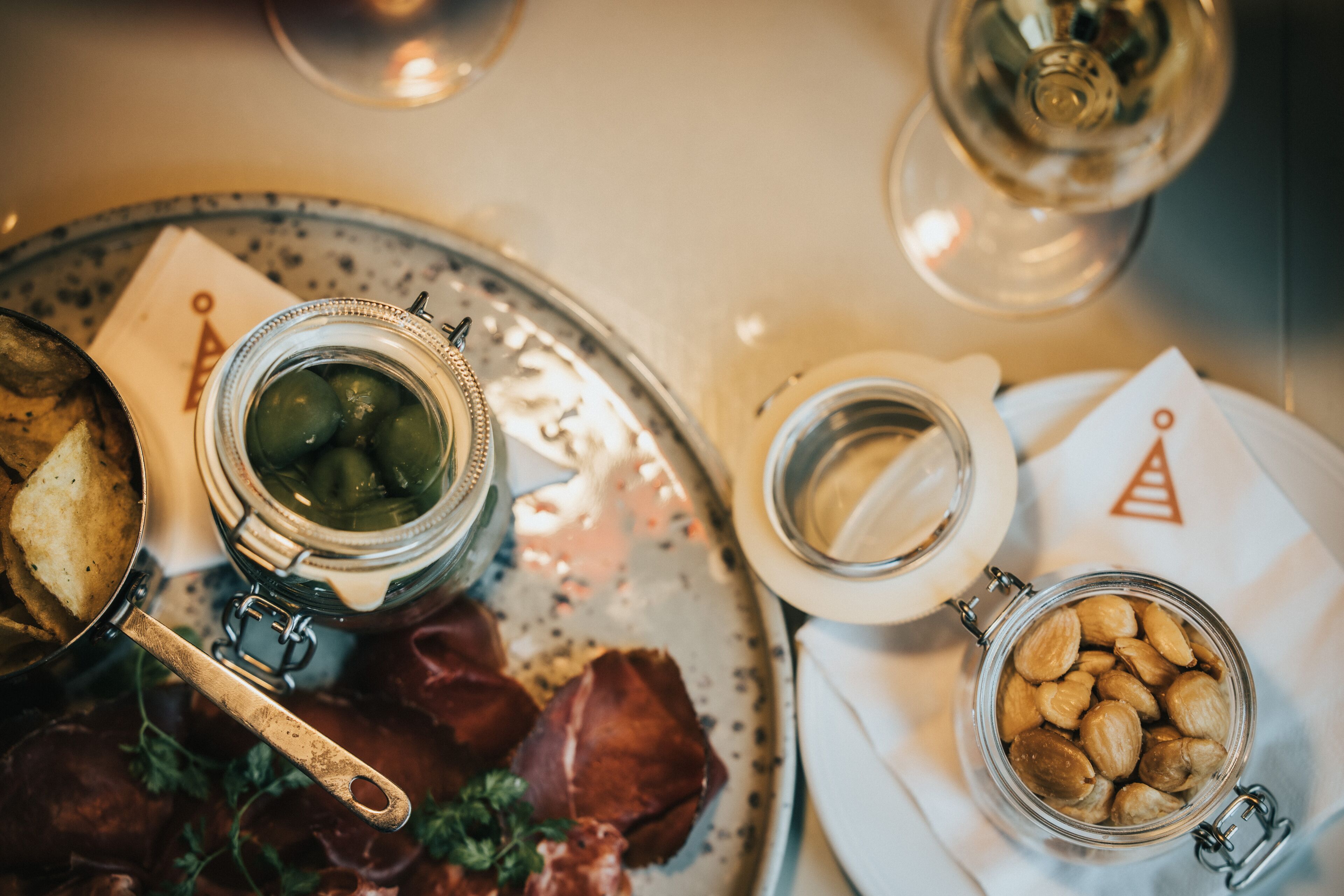 A dining table with a metal tray holding a jar of olives, a jar of almonds, and some bread.