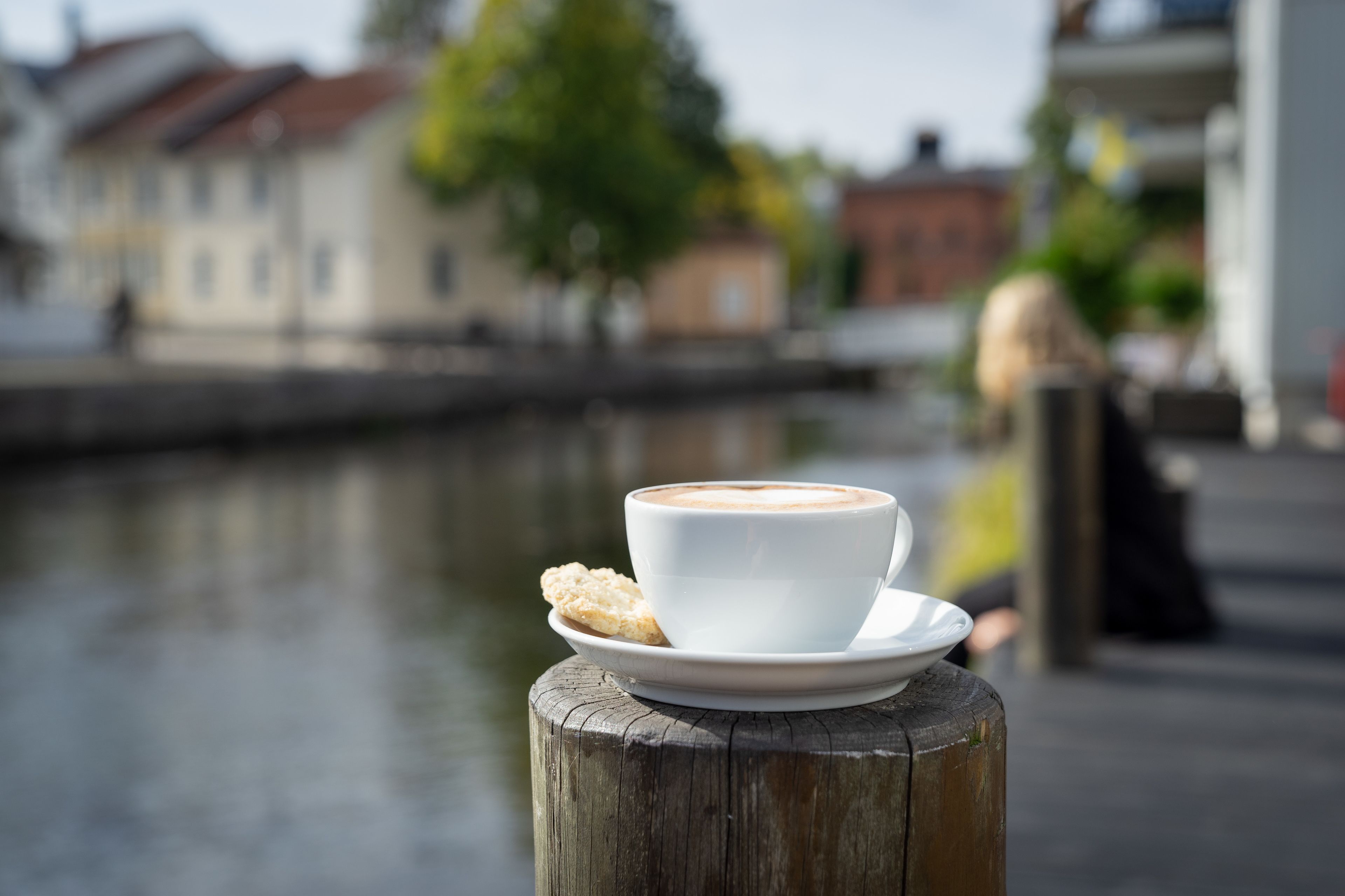 Una tazza di caffè su un pilastro di legno, con un biscotto su un piattino, con vista su un’acqua calma e edifici sullo sfondo.