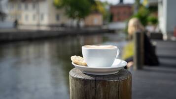 Una tazza di caffè su un pilastro di legno, con un biscotto su un piattino, con vista su un’acqua calma e edifici sullo sfondo.