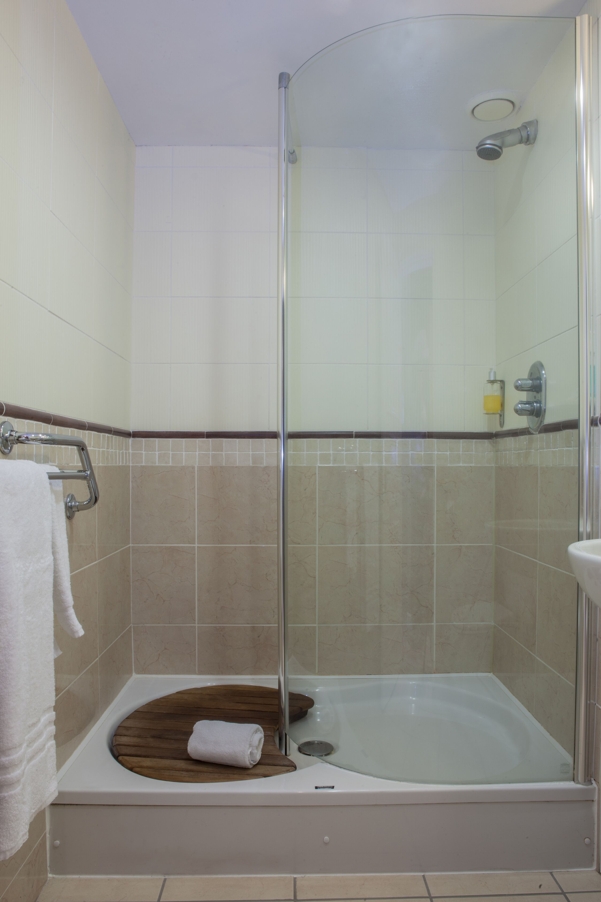 A walk-in shower with a glass door, a white bathtub with a wooden seat, and a white towel on the floor.