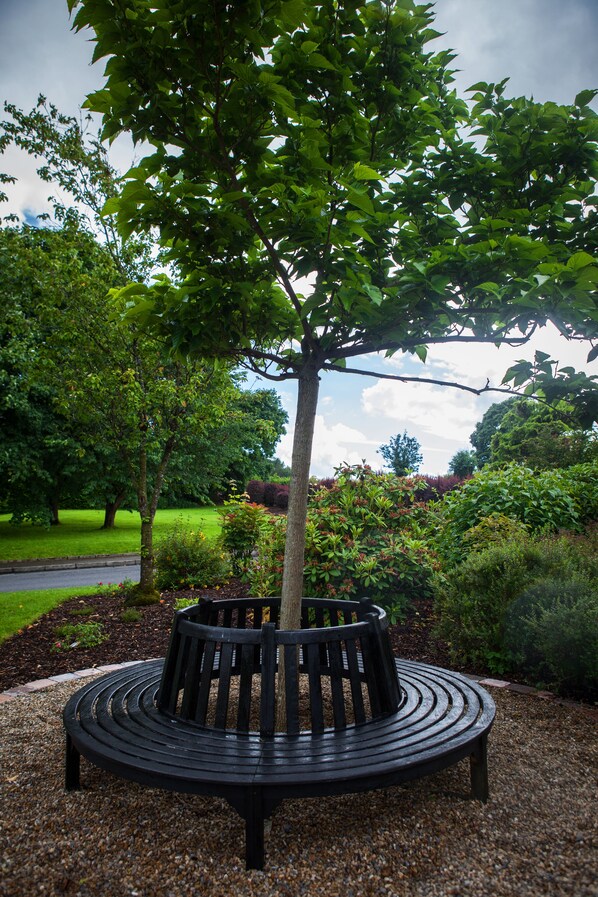 Un banco de madera circular con un tronco de árbol en el centro, rodeado de grava, en un parque con diferentes árboles y arbustos.