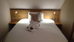 A neatly made bed with a white duvet, a folded towel, and a plush toy on the center. There are two bedside tables with lamps, and a wooden headboard. - Penny Farthing Hotel (Lyndhurst)
