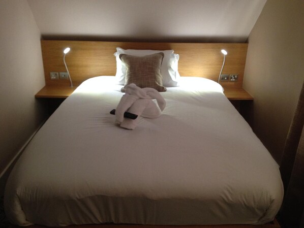 A neatly made bed with a white duvet, a folded towel, and a plush toy on the center. There are two bedside tables with lamps, and a wooden headboard.