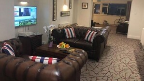 A living room with a leather sofa set, a coffee table with fruit, and a television on the left. - Penny Farthing Hotel (Lyndhurst)