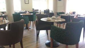 A dining area with round tables, green upholstered chairs, and a clock on the wall. - Penny Farthing Hotel (Lyndhurst)