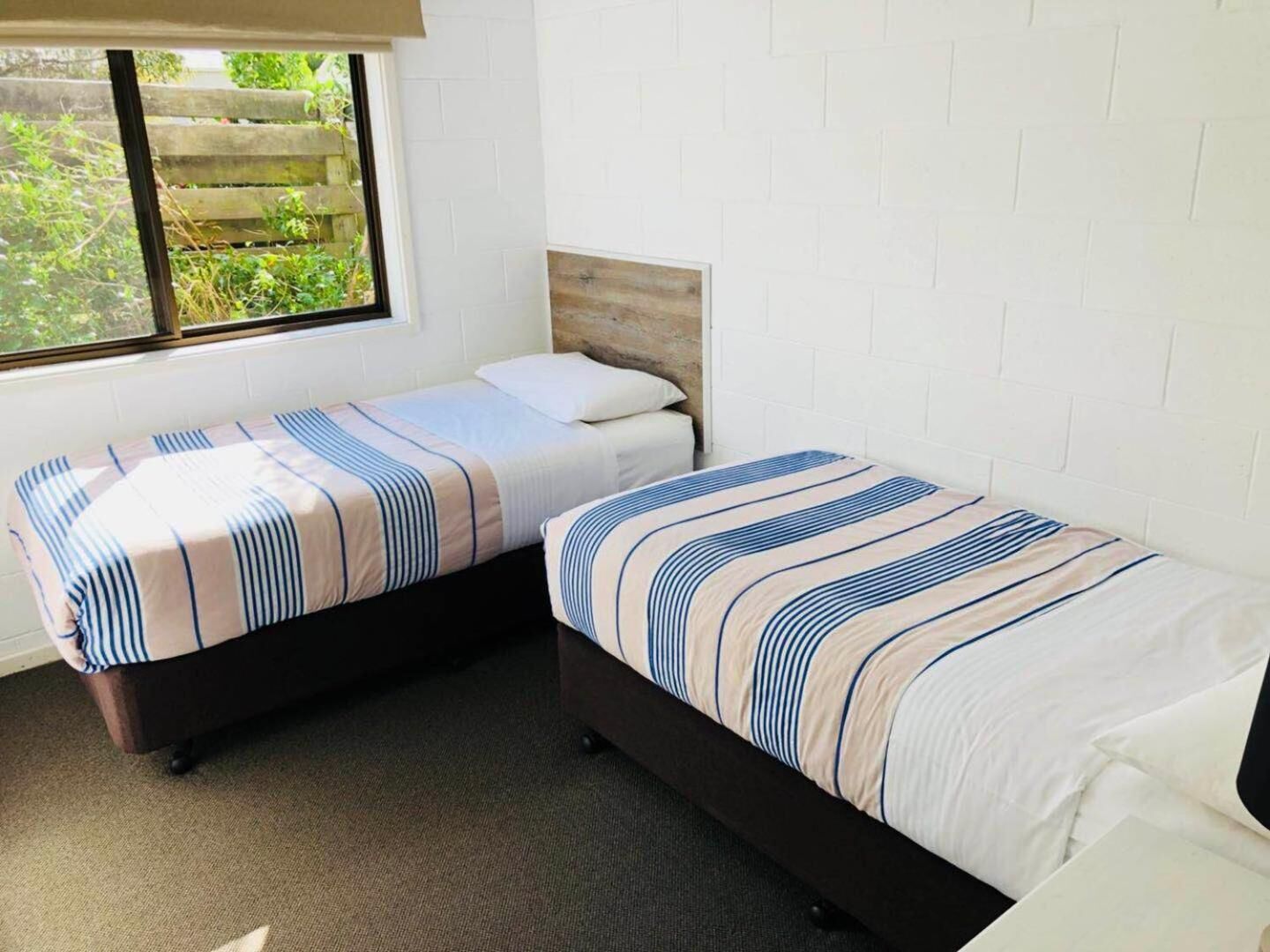 A small, clean room with two single beds, a window with a view of greenery, and white brick walls.