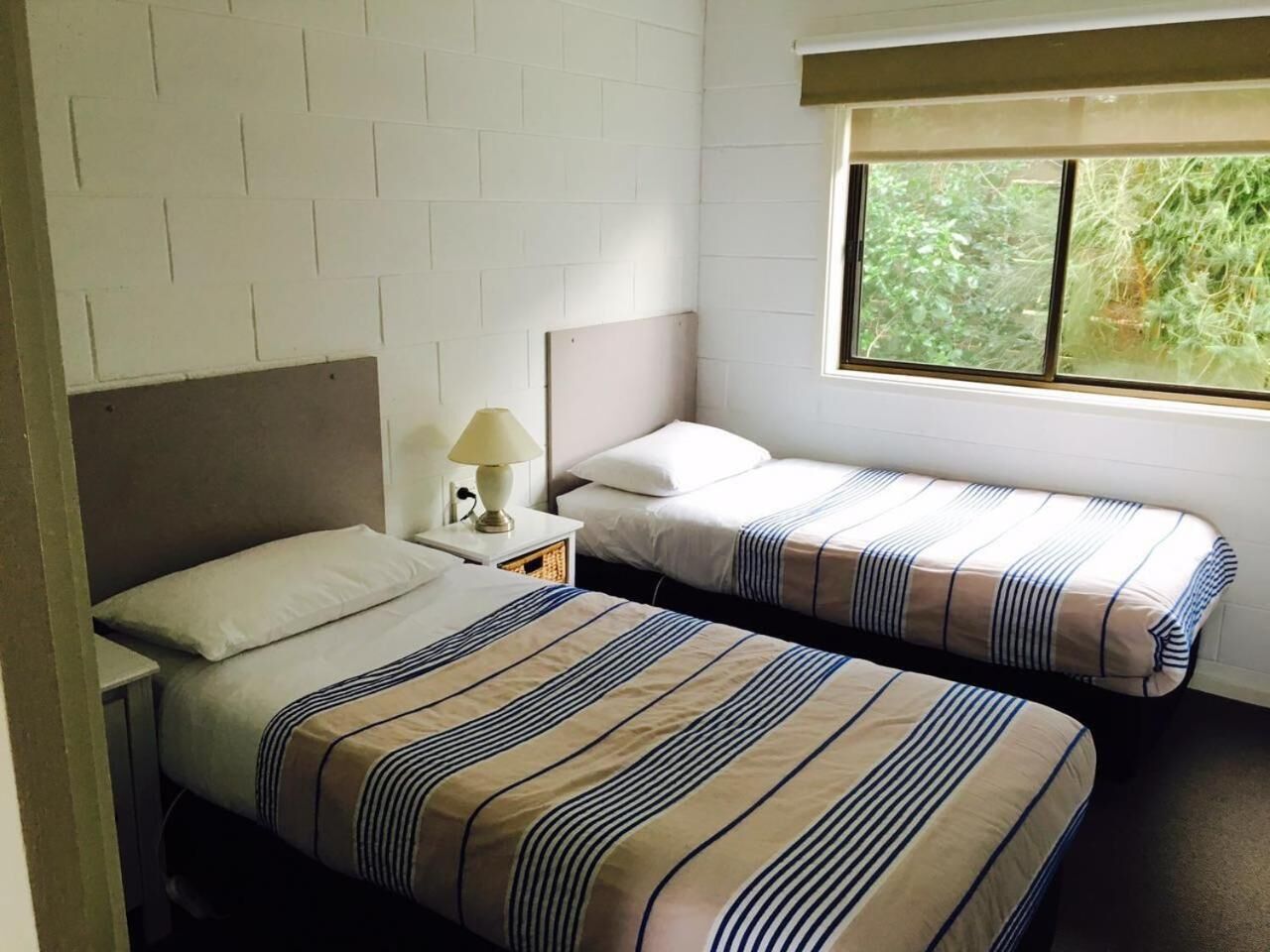 A room with two beds, each with striped bedding, a nightstand with a lamp, and a window with a view of greenery.