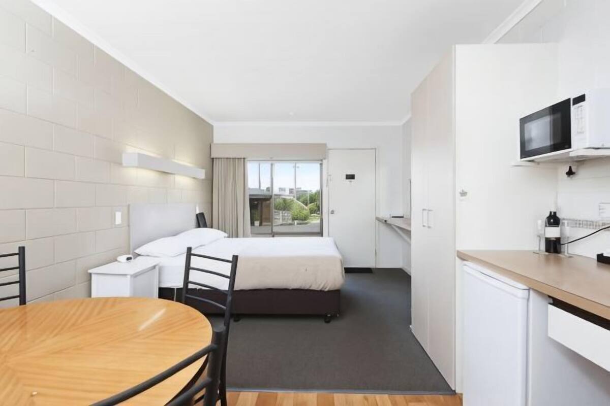 A hotel room with a bed, bedside table, desk, chair, and a small kitchenette.
