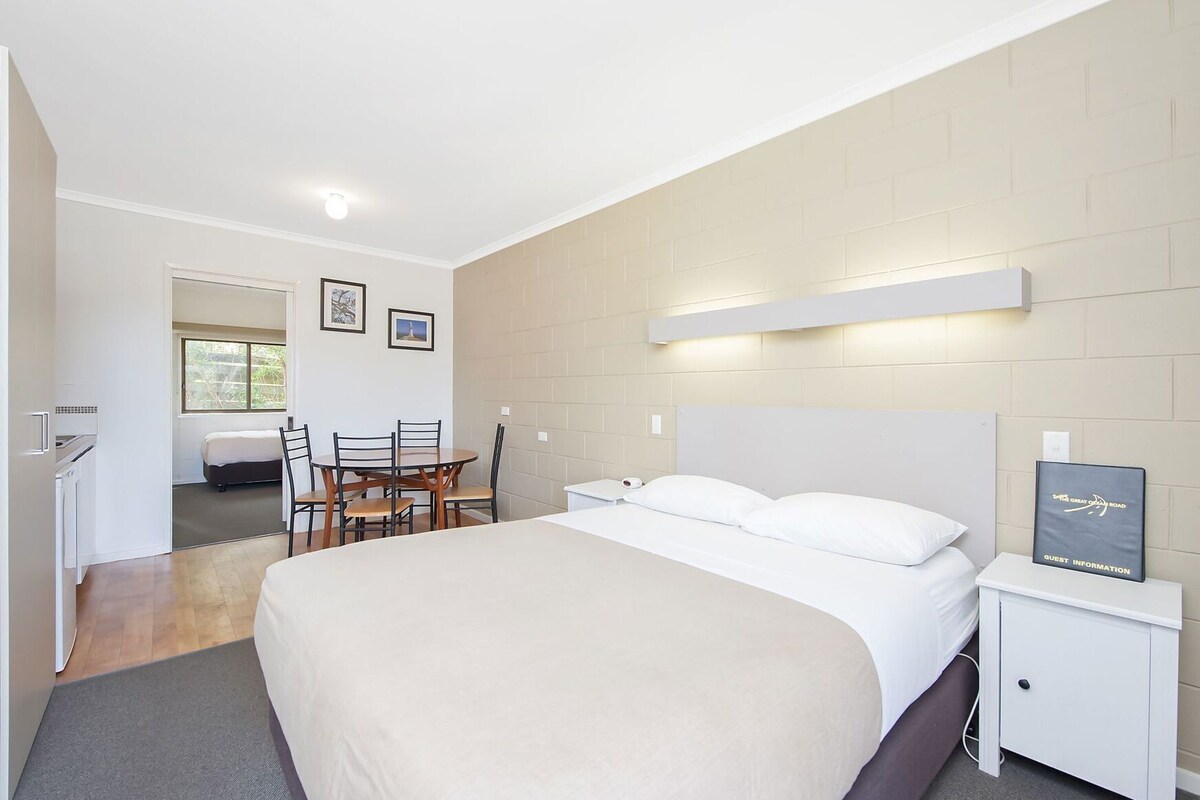 A modern hotel room with a large bed, bedside table, and a desk with chairs. There is a window with a view of greenery outside.