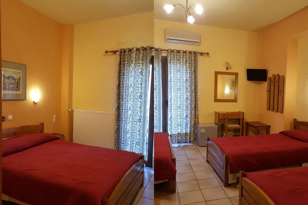 Habitación de hotel con dos camas individuales, una mesita de noche de madera, una silla, una pequeña mesa, un espejo, un televisor y una ventana con cortinas estampadas.