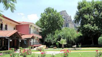 Un hôtel avec un toit recouvert de tuiles rouges, un jardin fleuri et un palmier, et une montagne en arrière-plan.