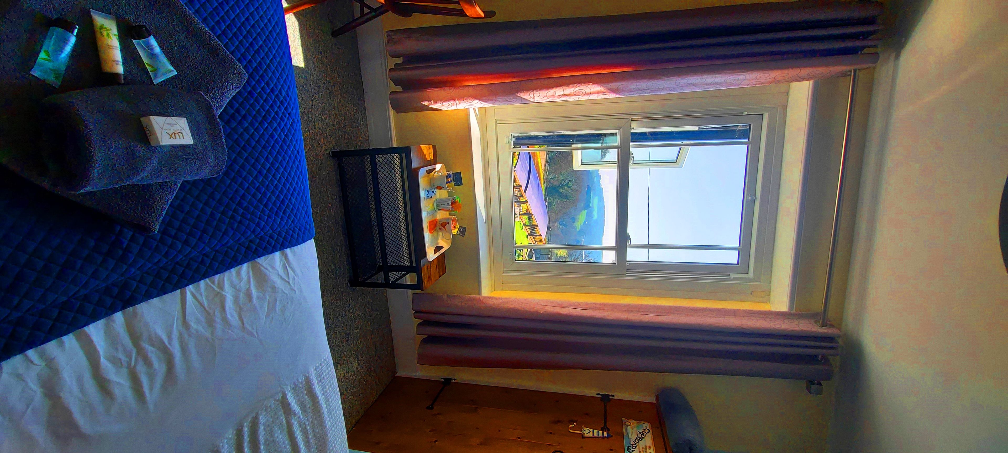 A room with a bed, a window with curtains, a small table with items, and a wall with a shelf.