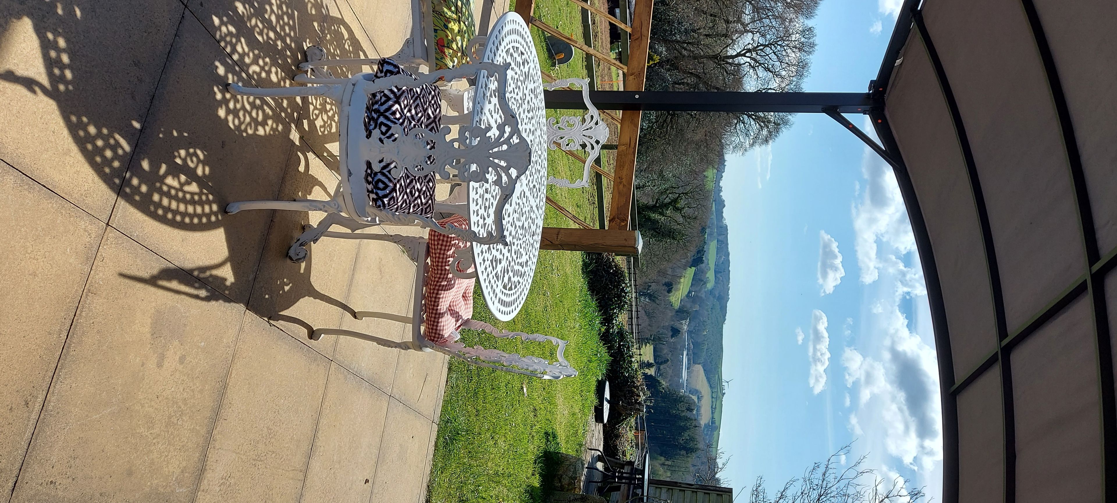 A patio area with a metal table and chairs, a patterned umbrella, and a view of a green landscape with a clear sky.