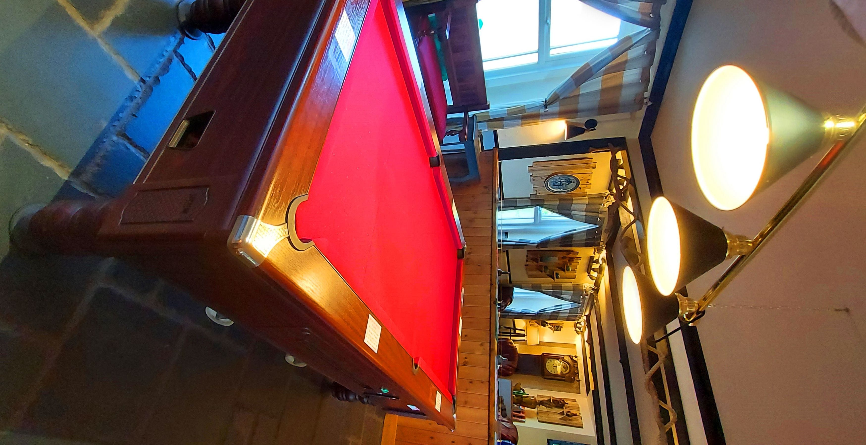 Una mesa de billar con superficie de fieltro rojo, estructura de madera y un reloj en la pared.