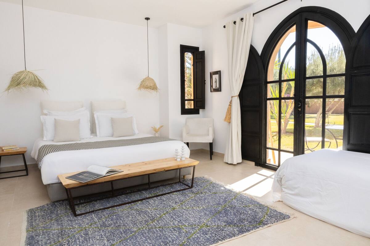 A bedroom with a bed, a bench, a reading area, a chair, a window with curtains, and a door.
