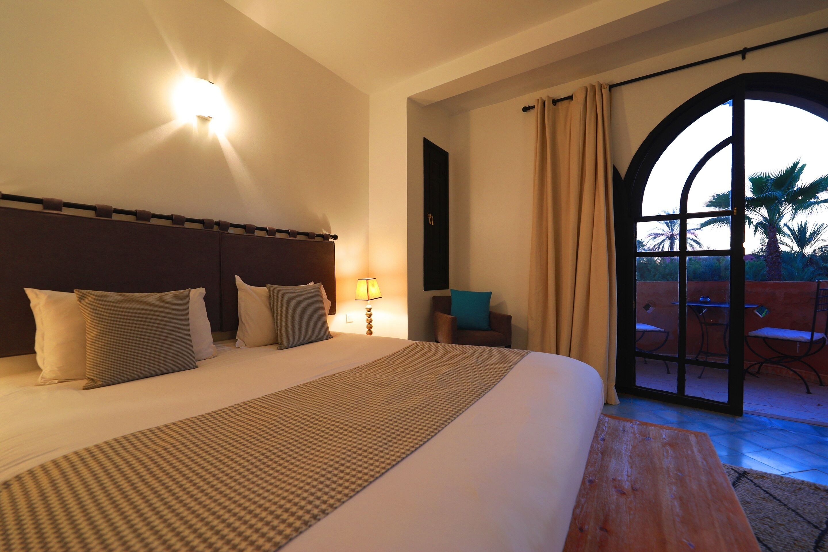 A hotel room with a large bed, a nightstand with a lamp, a chair, and a view of the outdoors through a glass door.