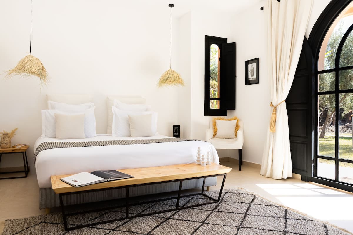 A bedroom with a bed, bedside tables, a reading bench, a window with curtains, and a view of greenery.