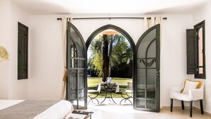 A bedroom with a bed, a wooden side table, a chair, and a view through a glass door to an outdoor seating area. -  Villa Al Assala Palmeraie - Piscine chauffée (Marrakech)