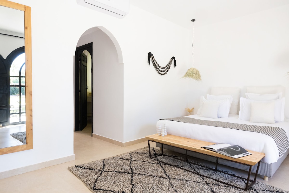 Un dormitorio con cama, un banco, un espejo y una lámpara colgante.