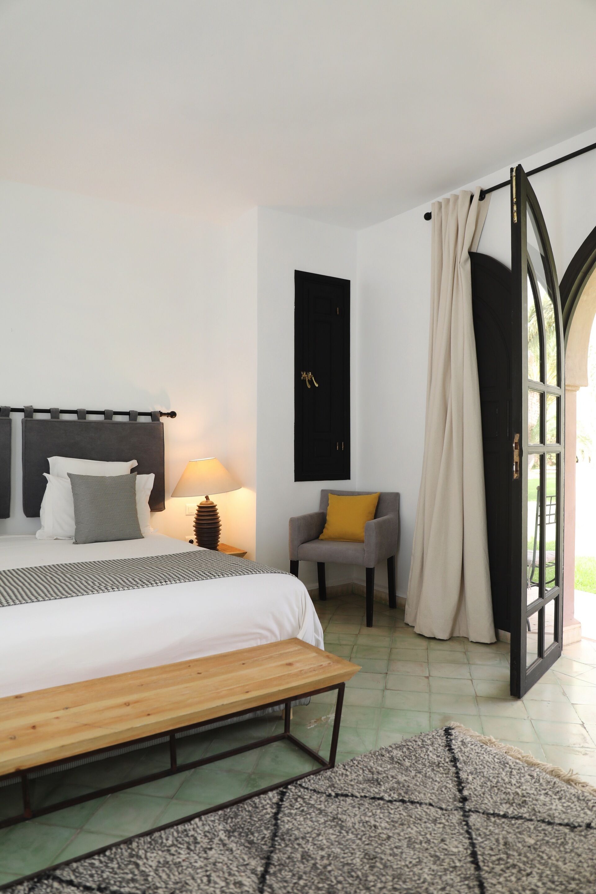 Ein Schlafzimmer mit Bett, Nachttisch, Lampe, Stuhl und Blick durch eine offene Tür.