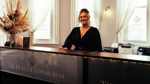 A reception desk with "The Duke of Edinburgh" branding, a vase with dried flowers, and a window with sheer curtains. - Duke Of Edinburgh Hotel (Barrow-in-Furness)
