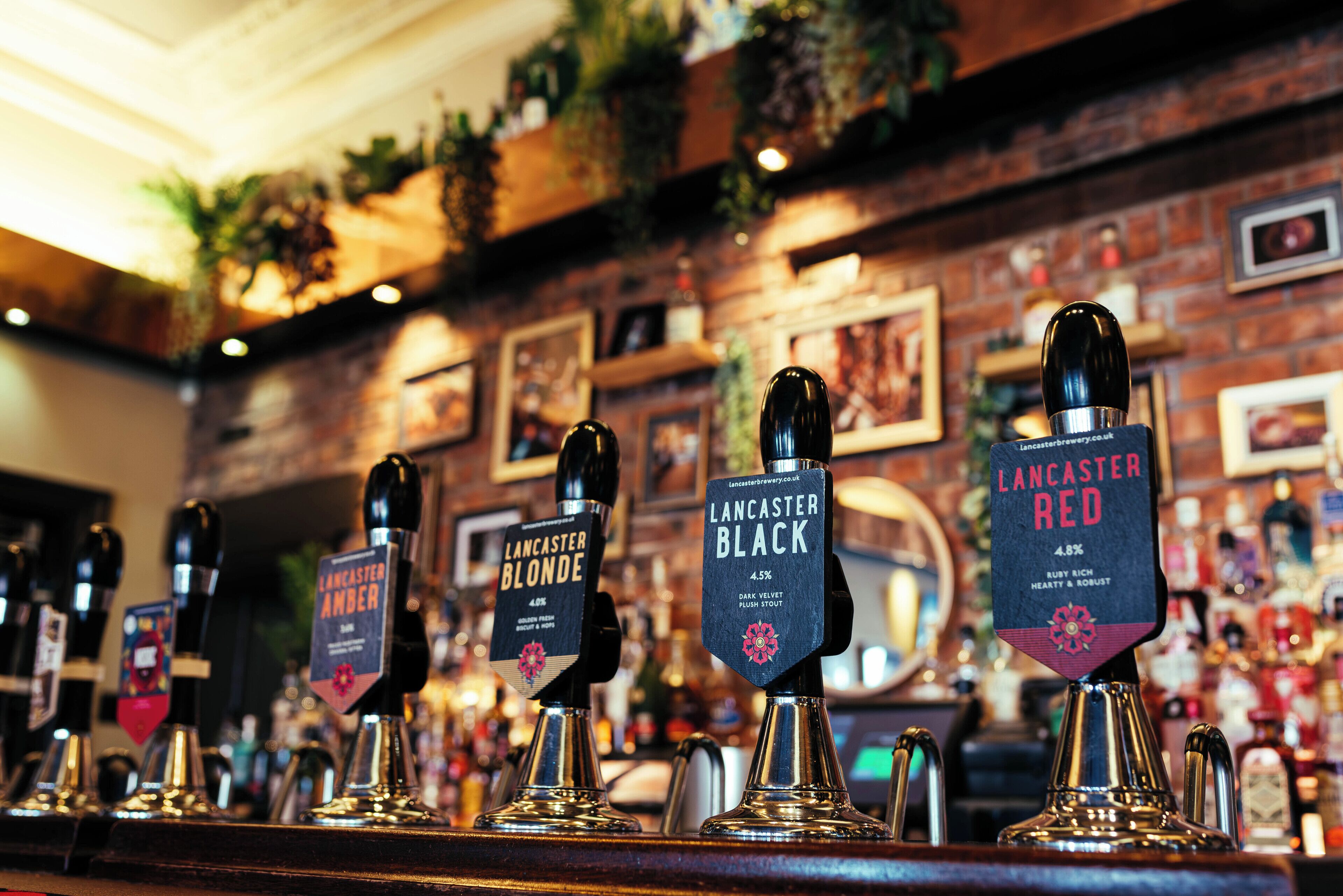 Een bar met meerdere tappunten, elk voorzien van een bordje met verschillende soorten bier, waaronder Lancaster Blonde en Lancaster Red.