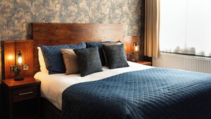 A neatly made bed with a dark blue quilt and multiple pillows, a wooden headboard, and two bedside lamps on wooden nightstands. - Duke Of Edinburgh Hotel (Barrow-in-Furness)